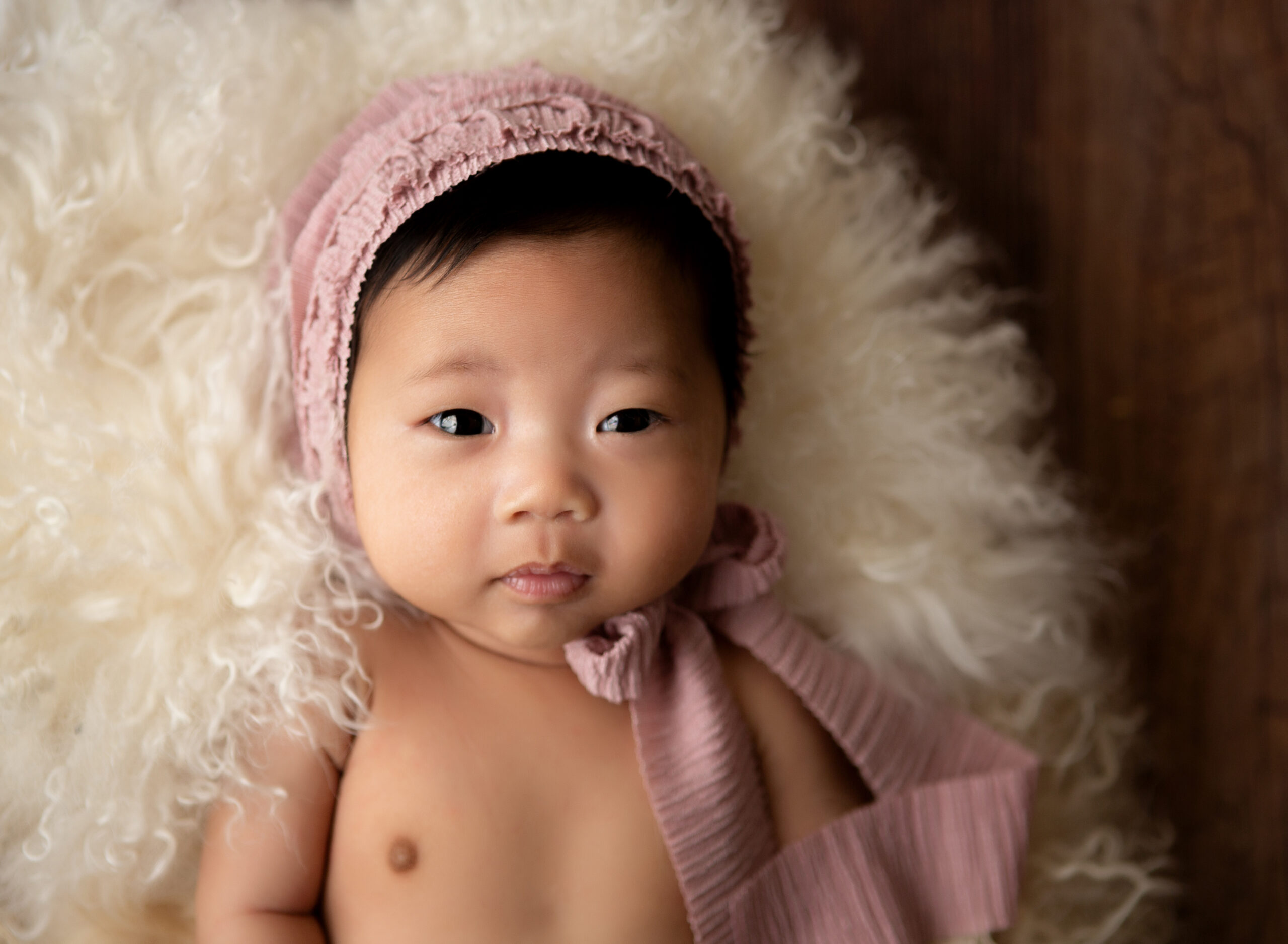 photograph of a 10 week old baby wearing a pink bonnet lying on a neutral fur looking into the camera